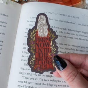 May include: A sticker featuring a woman with long white hair and a red dress, framed by a floral design. The text "FROM NOW UNTIL THE DARKNESS CLAIMS US" is displayed on the sticker. The sticker is placed on an open book.