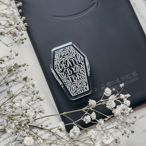 May include: A black e-reader with the Amazon logo, a coffin-shaped sticker that reads "DEATH BY TBR", and a book with white flowers. The e-reader is open on a book with text. Decorative gold spheres are in the background.