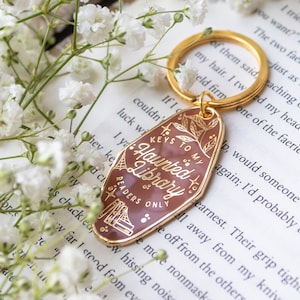 Puede incluir: Un llavero marr&oacute;n y dorado con el texto "Keys to my Haunted Library Readers Only". El llavero tiene forma de llave de hotel vintage y est&aacute; rodeado de flores blancas y un libro.