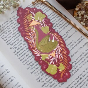 May include: A burgundy bookmark with a dragon illustration, gold sword-shaped bookmark, and a lit candle. The dragon design features green and gold accents. The bookmark is resting on an open book.