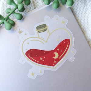 May include: A heart-shaped glass bottle filled with a red liquid and gold glitter. The bottle has a cork stopper and a crescent moon design.