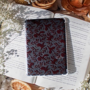 May include: A burgundy phone case with a floral pattern featuring small birds and leaves. The case is lying open on a book with a white background. The book has text on the pages, including the words "Sole of Thorns and Roses".
