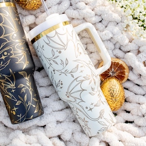 May include: Two stainless steel tumblers with a gold rim and a bat and vine design. The tumblers are on a white fuzzy blanket with gold decorative eggs and dried orange slices.