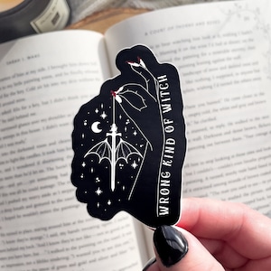 Puede incluir: Pegatina en blanco y negro con una mano sosteniendo una espada y un murciélago. La pegatina tiene una luna creciente y estrellas. El texto "Wrong Kind of Witch" está escrito verticalmente en la pegatina.
