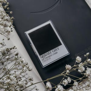 Puede incluir: Un lector electrónico negro con el logotipo de Amazon, una pequeña tarjeta enmarcada con el texto "MORALLY GREY MY FAVORITE COLOR #383838" y un libro con flores blancas. El lector está sobre un libro abierto.