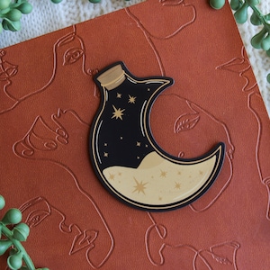 Puede incluir: Una botella con forma de media luna llena de un líquido brillante y estrellas doradas. La botella es negra con una tapa dorada y está sobre una superficie de cuero marrón.