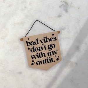 May include: A brown banner with the text "bad vibes don't go with my outfit." The banner has a black border and stars on each side. The banner is hanging from a string.