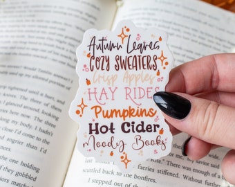 May include: A white sticker with orange, pink, and brown text that reads "Autumn Leaves, Cozy Sweaters, Crisp Apples, Hay Rides, Pumpkins, Hot Cider, Moody Books".