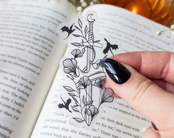 May include: A clear sticker with a black line drawing of a sword, crescent moon, and bats. The sword is surrounded by leaves and flowers. The sticker is on a book.