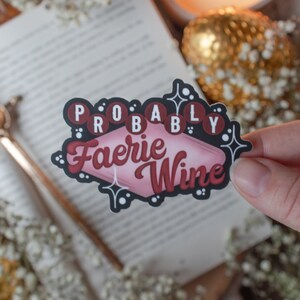 Puede incluir: Una pegatina rosa y negra con el texto "Probably Faerie Wine" en una fuente retro. La pegatina está rodeada de purpurina blanca y estrellas.