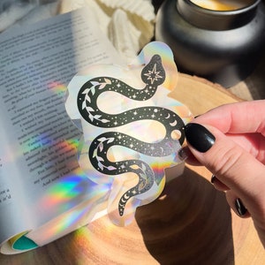 May include: A holographic sticker featuring a black snake with stars and crescent moons on a white background. The snake is curled around itself and has leaves growing from its body.