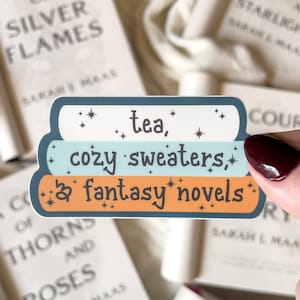 May include: A sticker with the text "tea, cozy sweaters, & fantasy novels" in a blue, light blue, and orange color scheme. The sticker is decorated with small white stars.