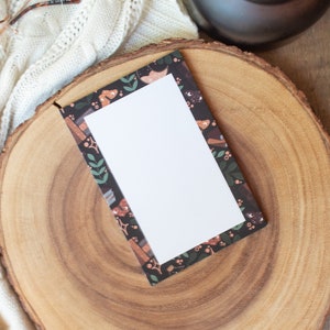 May include: A blank white card with a dark floral border sits on a wooden slice. The card is surrounded by a white knitted blanket and a black candle.