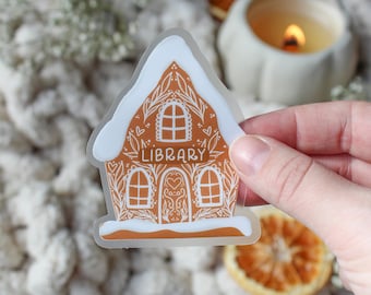 May include: A clear sticker with a white gingerbread house design. The house has a brown roof and white trim. The words "LIBRARY" are written on the front of the house.