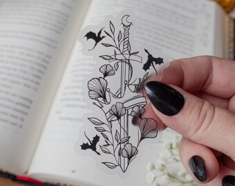May include: A clear sticker with a black line drawing of a sword, a crown, and three dragons. The sword is surrounded by floral designs.