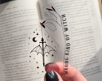 May include: A clear sticker with a black and white illustration of a hand holding a sword with wings. The background is a starry night sky with a crescent moon. The text "WRONG KIND OF WITCH" is written vertically on the right side of the sticker.