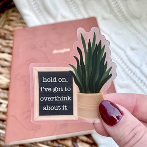 May include: A clear sticker with a black chalkboard background and white text that reads "hold on, i've got to overthink about it." A green plant in a brown pot is on the right side of the sticker.