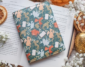 Puede incluir: Una cubierta de libro de color verde azulado con un patrón de dragones, libros y follaje en tonos naranja, blanco y beige. El libro está abierto, revelando el título "HORNS AND ROSES". La escena está adornada con flores y adornos dorados.