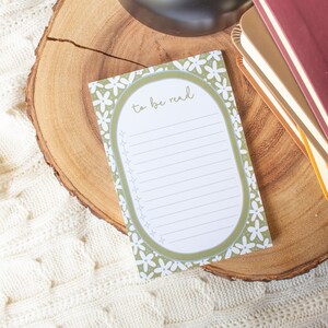May include: A green and white floral notepad with the text "to be read" on the front. The notepad is on a wooden surface with a stack of books and a candle.