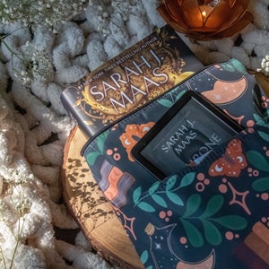 May include: A dark blue book sleeve with a floral pattern featuring moons, stars, and butterflies. The sleeve has a zipper closure and is holding a book titled "A Court of Thorns and Roses" by Sarah J. Maas.