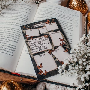 K&ouml;nnte beinhalten: Eine Notizkarte f&uuml;r B&uuml;cher mit einem Schmetterlingsdesign liegt auf einem offenen Buch. Die Karte hat Abschnitte f&uuml;r Titel, Autor, Genre, Lieblingszitat und Notizen. Wei&szlig;e Blumen und ein goldenes Ei umgeben die Karte.