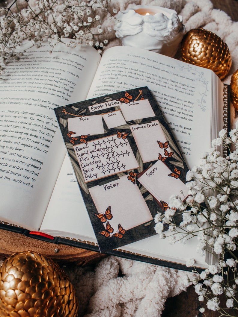 K&ouml;nnte beinhalten: Eine Notizkarte f&uuml;r B&uuml;cher mit einem Schmetterlingsdesign liegt auf einem offenen Buch. Die Karte hat Abschnitte f&uuml;r Titel, Autor, Genre, Lieblingszitat und Notizen. Wei&szlig;e Blumen und ein goldenes Ei umgeben die Karte.