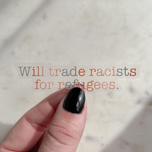 May include: A clear sticker with the text "Will trade racists for refugees." The words "racists" and "refugees" are in red and orange, respectively. The sticker is held by a hand with a black painted nail.