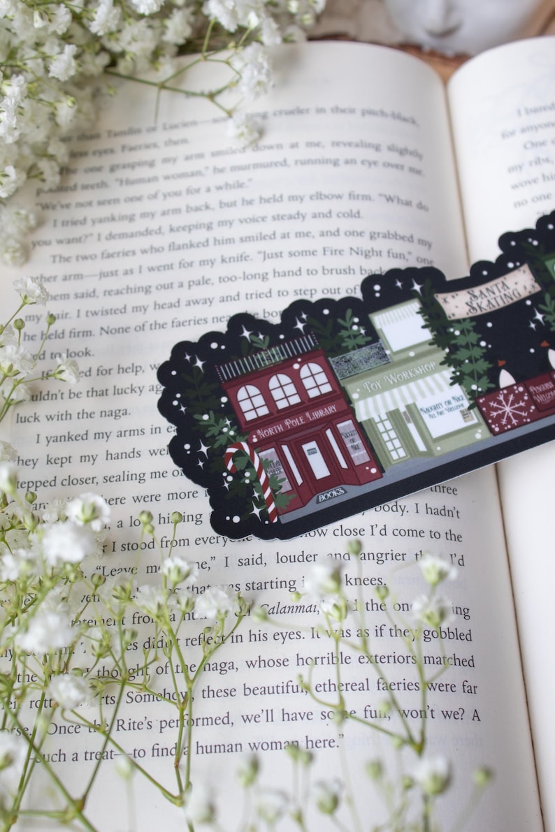 May include: A black and white sticker with a Christmas theme. The sticker features a red building with the words "North Pole Library" and "Books" on it. There are also two other buildings, one with the words "Toy Workshop" and the other with the words "Santa Skating".