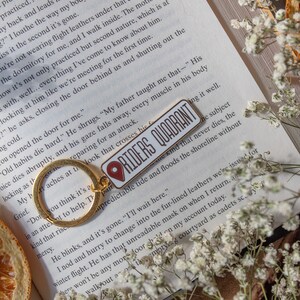 Op de afbeelding: Een gouden sleutelhanger met een rode en witte emaille pin waarop "Riders Quadrant" staat. De pin is bevestigd aan een gouden sleutelring. De sleutelhanger ligt op een open boek met witte bloemen.