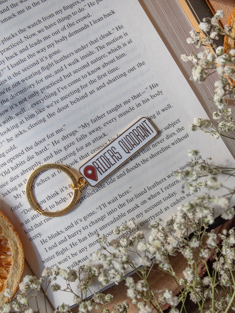 Op de afbeelding: Een gouden sleutelhanger met een rode en witte emaille pin waarop "Riders Quadrant" staat. De pin is bevestigd aan een gouden sleutelring. De sleutelhanger ligt op een open boek met witte bloemen.