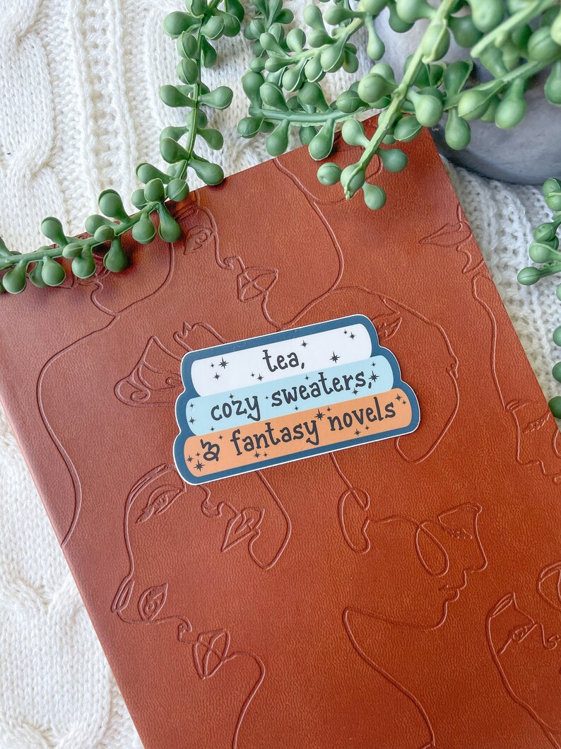 May include: A sticker with a blue, white, and light blue color scheme. The sticker has the text "tea, cozy sweaters, & fantasy novels" with stars around the text.