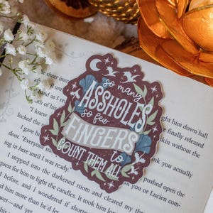 May include: A burgundy sticker with the text "so many assholes so few fingers to count them all" in a decorative font. The sticker has floral and star accents and is placed on an open book.