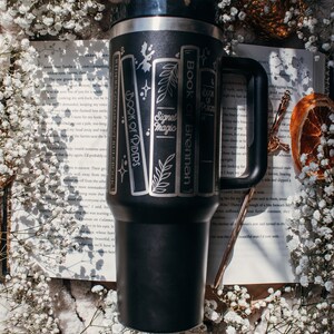 May include: A black stainless steel travel mug with a white design of books with titles "Book of Brennan", "Signet of Magic", and "Book of Riders". The mug is sitting on a white book with text.