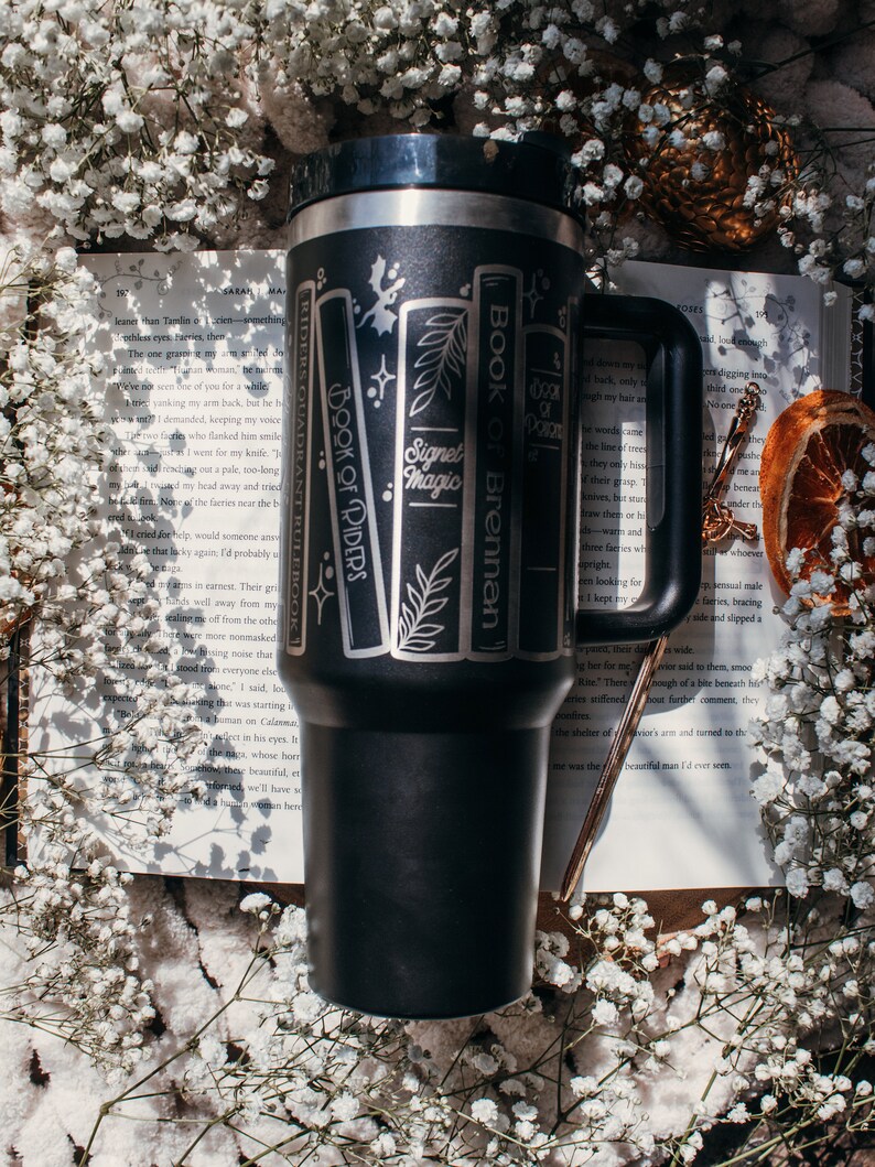 May include: A black stainless steel travel mug with a white design of books with titles "Book of Brennan", "Signet of Magic", and "Book of Riders". The mug is sitting on a white book with text.