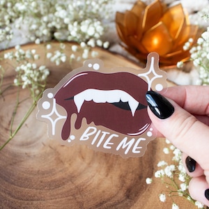 May include: A transparent sticker featuring a pair of vampire lips in shades of burgundy and white, with the text "BITE ME". The sticker has a glossy finish and is surrounded by decorative starbursts.