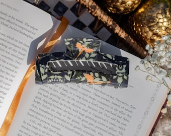 May include: A rectangular hair claw clip featuring a dark background with a repeating pattern of light green geometric lines and orange bird silhouettes. The clip is positioned on an open book page with visible printed text and surrounding greenery.