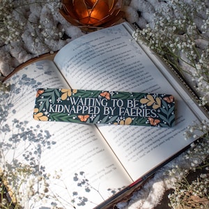 May include: A green and yellow bookmark with the text "Waiting to be Kidnapped by Fairies." The bookmark is decorated with flowers, butterflies, and stars. It is lying open on a book.