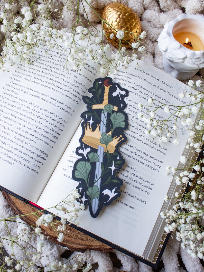 May include: A black and green bookmark with a sword, crown, and ginkgo leaves. The bookmark has a dark background with white stars and the text "A Court of Thorns and Roses".