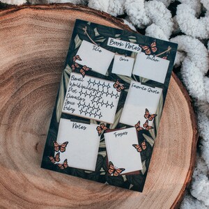 K&ouml;nnte beinhalten: Ein dekoratives Notizblatt f&uuml;r B&uuml;cher mit einem dunkelgr&uuml;nen Hintergrund und wei&szlig;en K&auml;stchen zum Schreiben. Das Blatt tr&auml;gt oben die Worte "Book Notes" und ist mit orangefarbenen Schmetterlingen verziert. Das Blatt liegt auf einer Holzoberfl&auml;che.