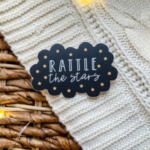 Peut inclure: Un autocollant noir et or en forme de nuage avec le texte "RATTLE the stars" en lettres blanches.