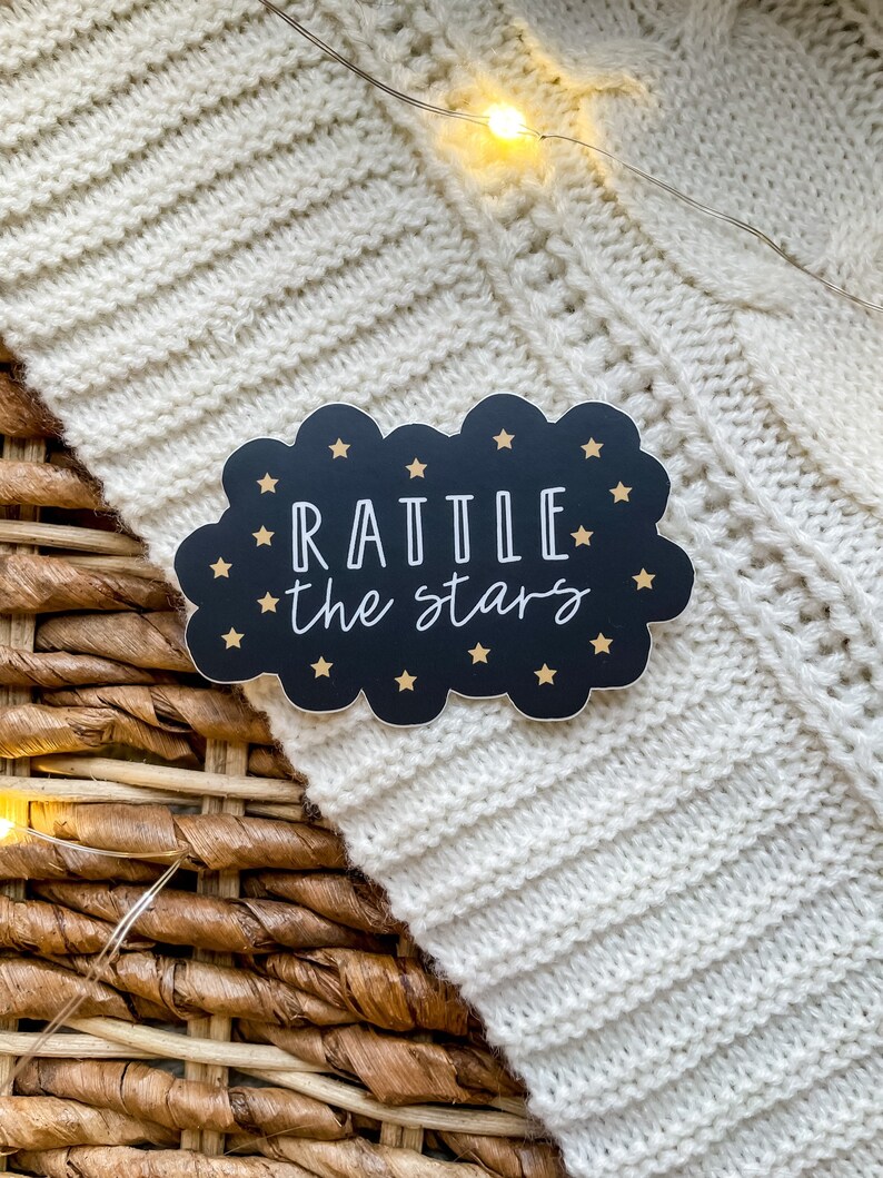 Peut inclure: Un autocollant noir et or en forme de nuage avec le texte "RATTLE the stars" en lettres blanches.