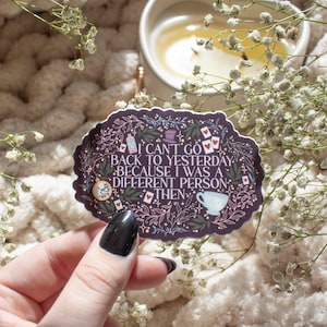 May include: A sticker with the quote "I can't go back to yesterday because I was a different person then." The sticker has a dark purple background with white text and Alice in Wonderland themed illustrations. The sticker is held in a hand.