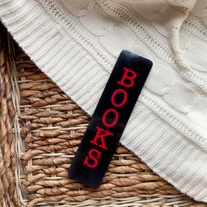 May include: A black bookmark with the word "BOOKS" in red letters. The bookmark is lying on a woven wicker surface and a white knitted sweater.