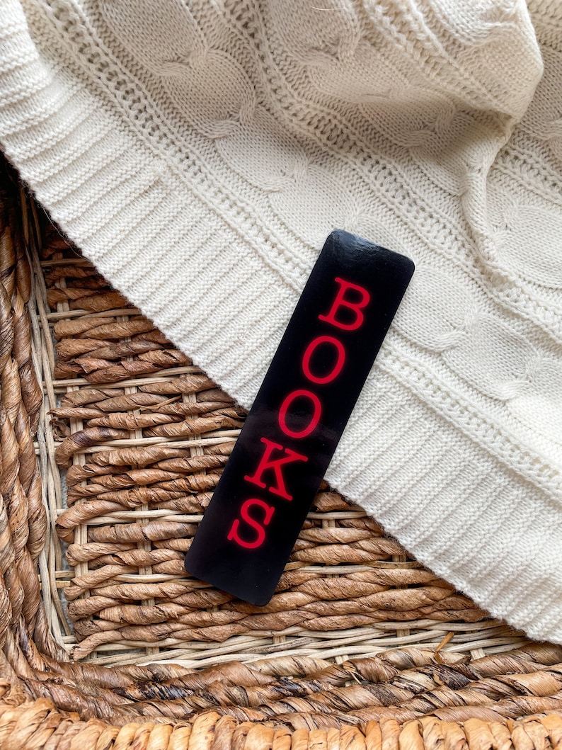 May include: A black bookmark with the word "BOOKS" in red letters. The bookmark is lying on a woven wicker surface and a white knitted sweater.
