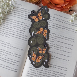 May include: A decorative bookmark featuring a design of orange and black monarch butterflies with green leaves. The bookmark is a vertical rectangle with a black background and white star accents. It is placed on an open book.