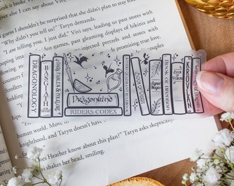 May include: A clear bookmark featuring a black and white illustration of stacked books, a potion bottle, and bats. The bookmark includes the text "Dragonkind Riders Codex" and other book titles. The bookmark is placed on an open book.