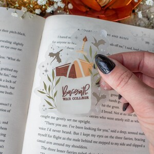 May include: A transparent sticker with a design of books, a sword, and the words "Bassiath War College." The sticker is placed over an open book. The design includes leaves and stars. The sticker is held by a hand.