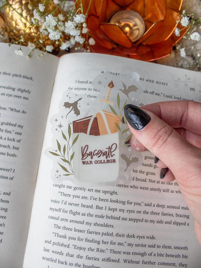 May include: A transparent sticker with a design of books, a sword, and the words "Bassiath War College." The sticker is placed over an open book. The design includes leaves and stars. The sticker is held by a hand.