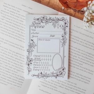 Puede incluir: Tarjeta de reseña de libro en blanco y negro con elementos de diseño floral y espada. La tarjeta tiene espacios para título, autor, género, notas y escalas de calificación para general, construcción del mundo, trama, personajes, picante y final.