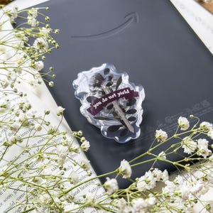 May include: A black e-reader with a clear resin embellishment. The resin piece contains dried flowers and a burgundy ribbon with the text "you do not yield." White flowers and a book are in the background.
